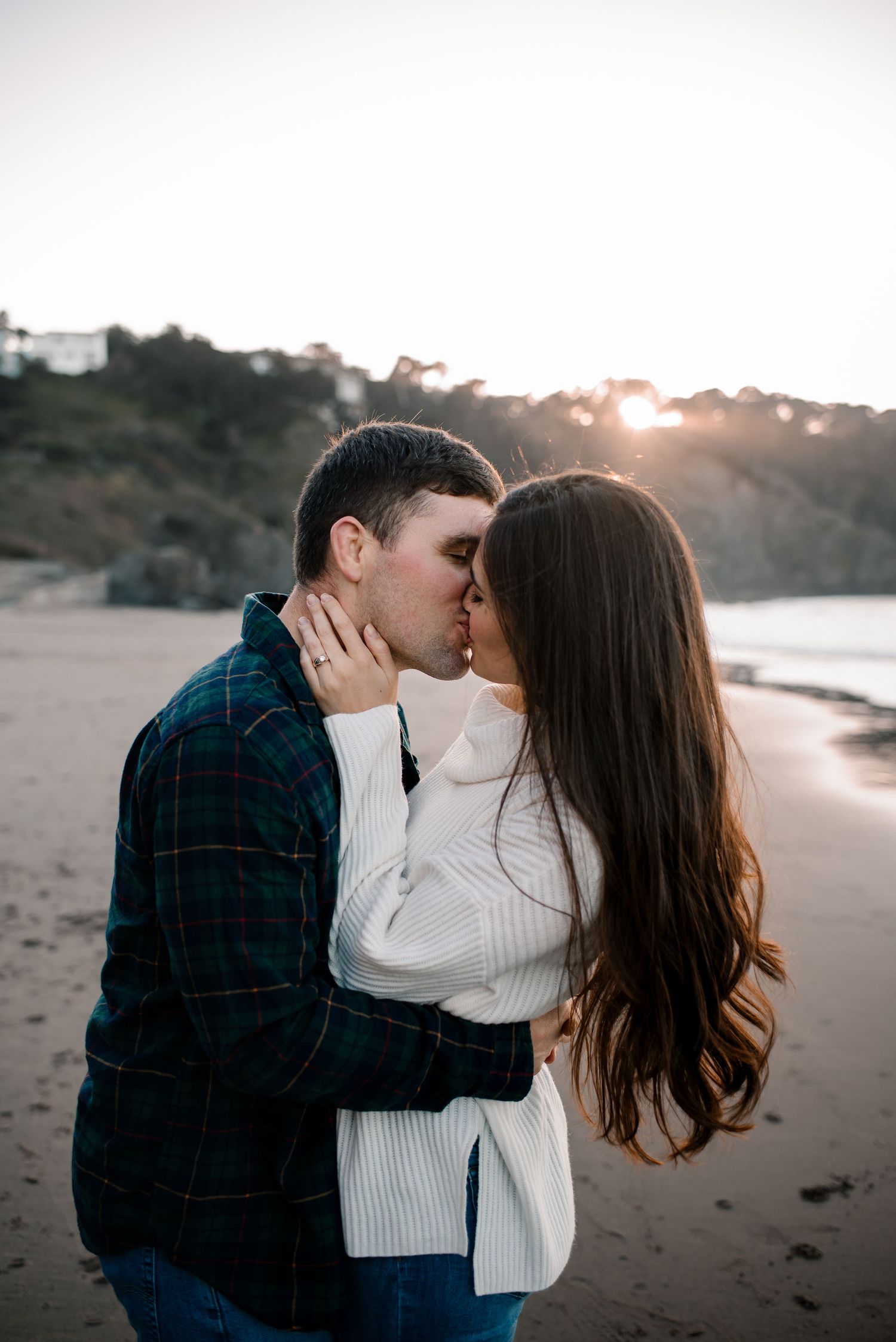 An intimate kiss shared between a couple on a beach with the sun setting behind coastal cliffs.