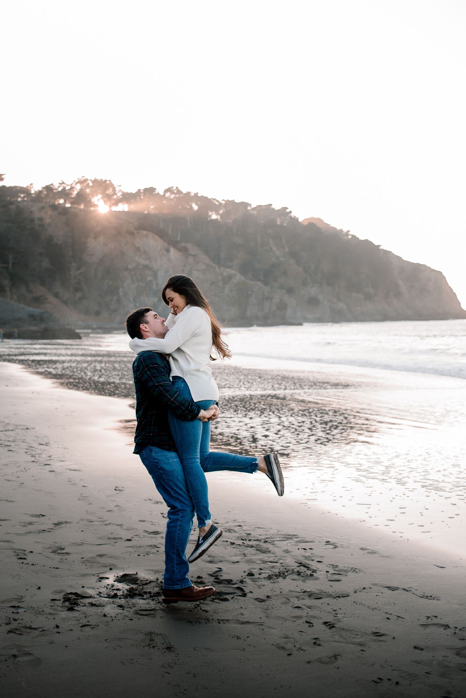 A romantic couple embracing on a beach at sunset with cliffs in the background.