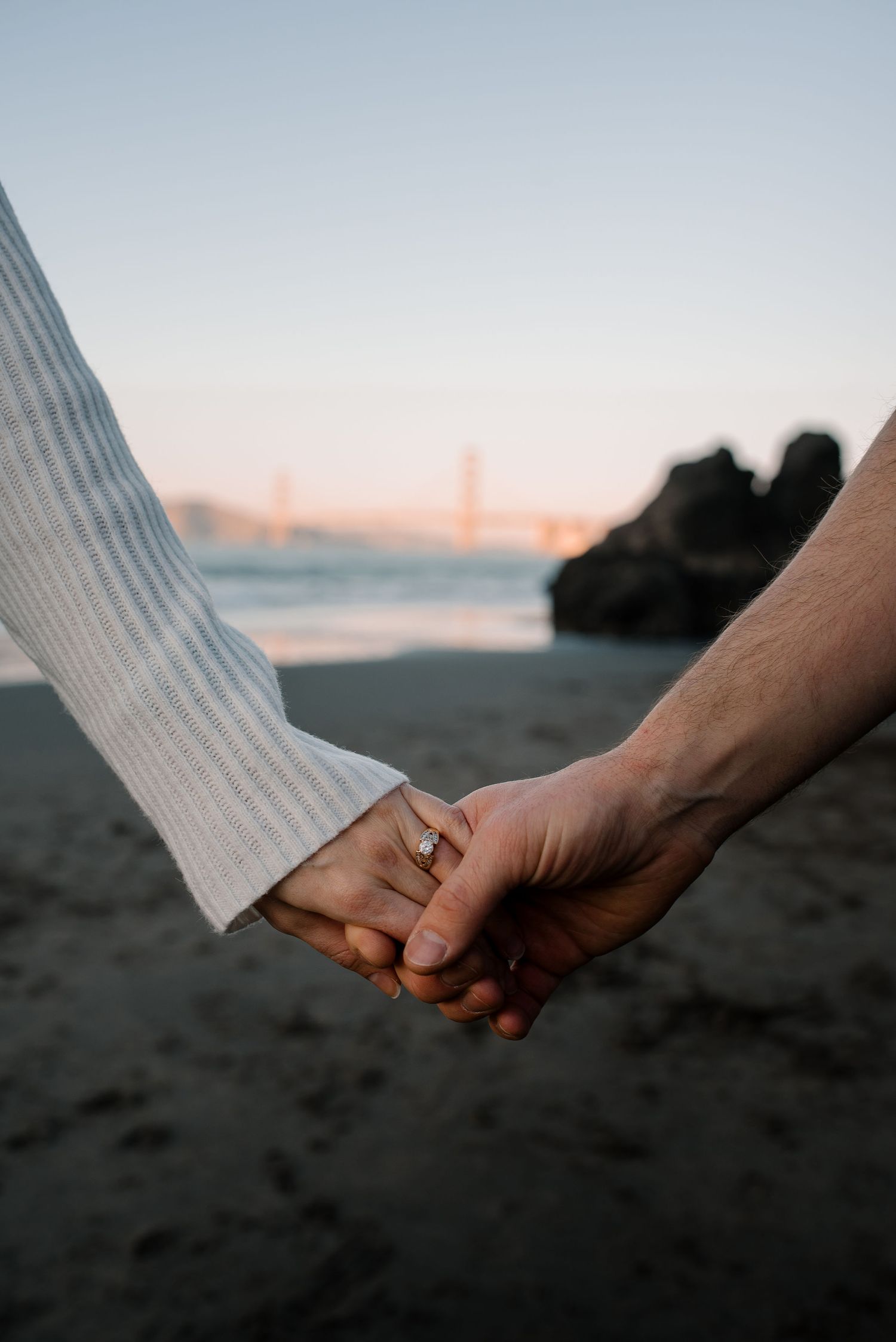 Close-up of two hands clasped together on a dark sandy beach at sunset.