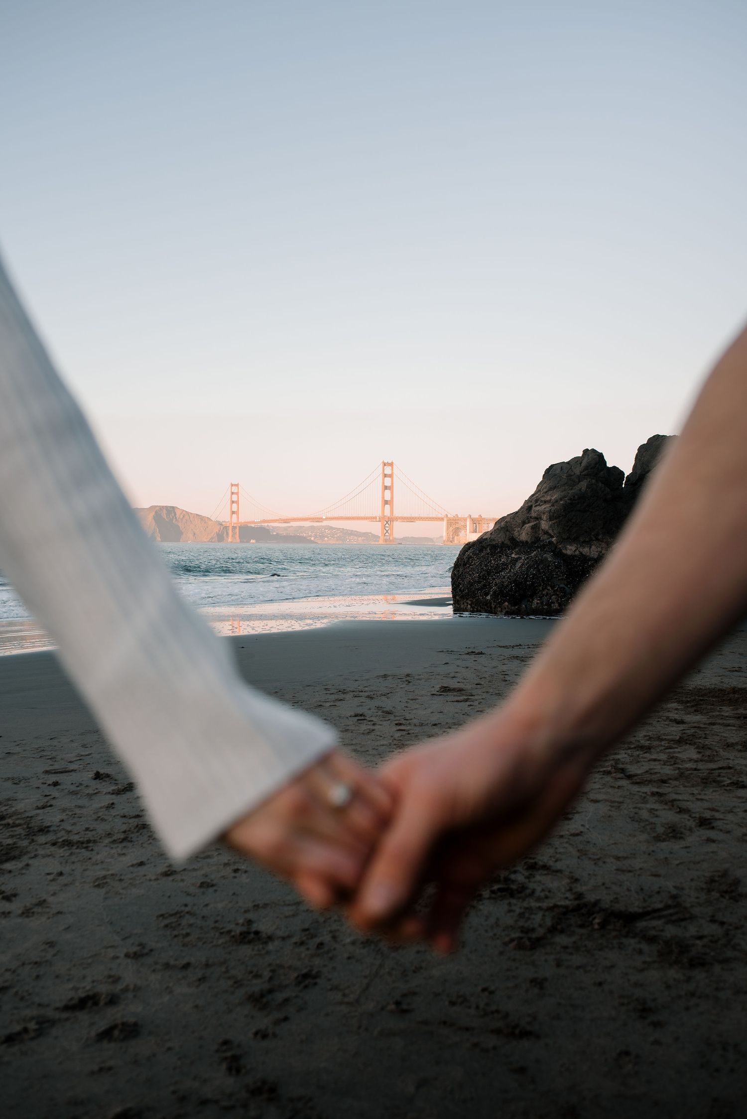 Two people hold hands with the Golden Gate Bridge visible in the distance across the bay.