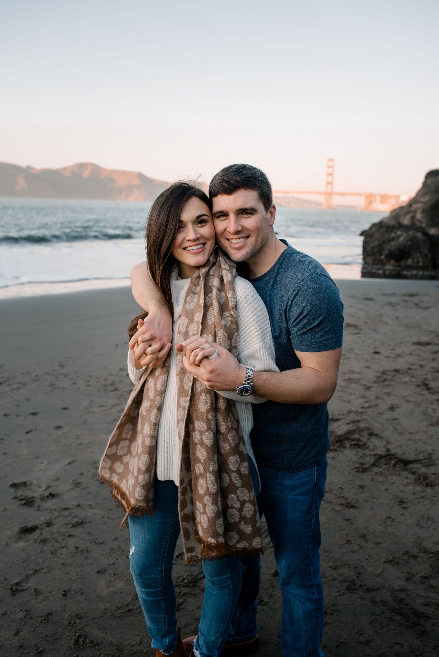 A couple embraces on a beach at sunset with San Francisco Bay in the background.