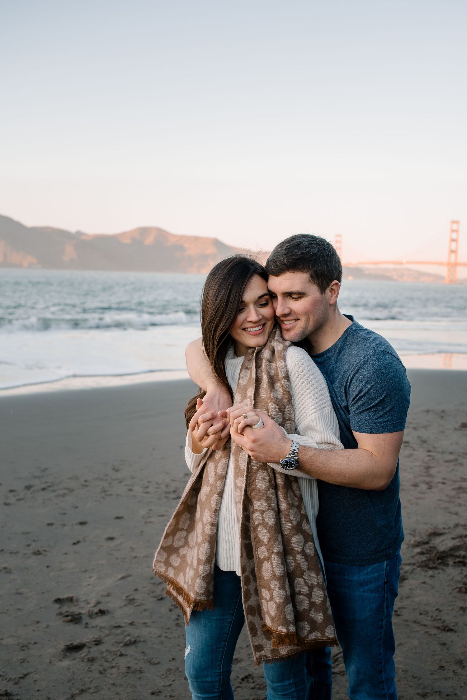 A couple shares an intimate moment on a beach with the Golden Gate Bridge visible in the background.