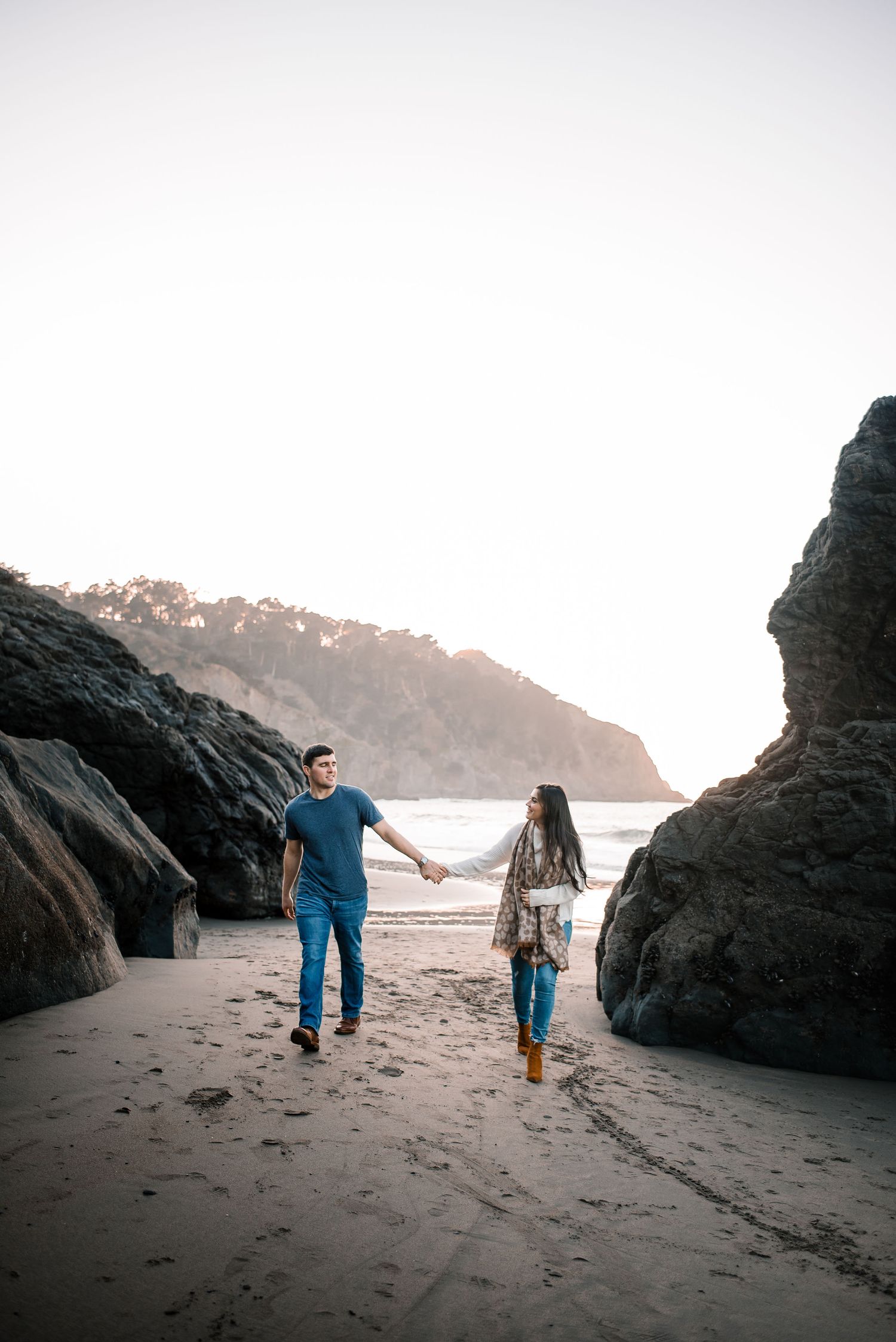 A couple holds hands while walking between large rock formations on a misty beach.
