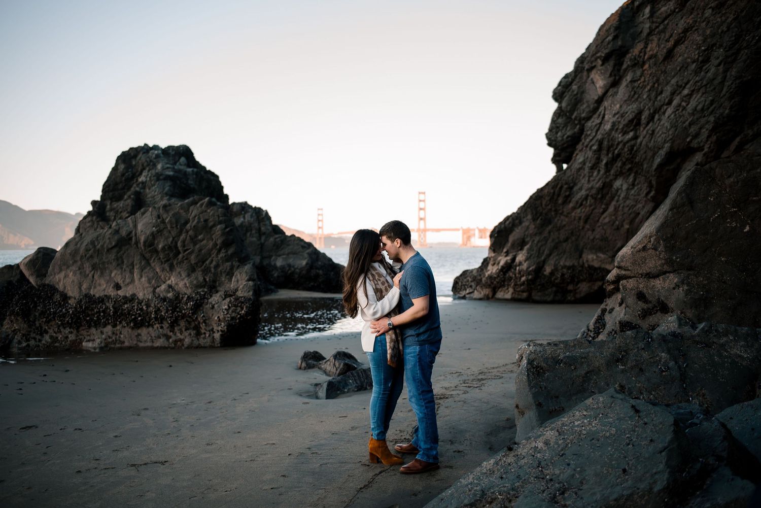 Two people share a romantic moment on a dark sandy beach near rocky cliffs.