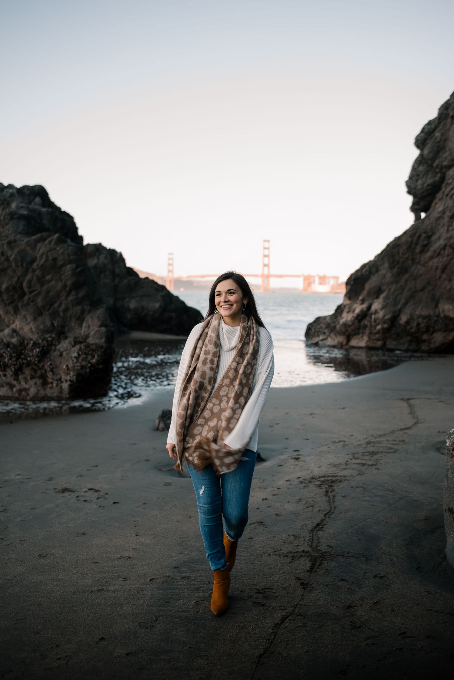 A couple walks along a rocky beach with the Golden Gate Bridge visible in the distance at sunset.