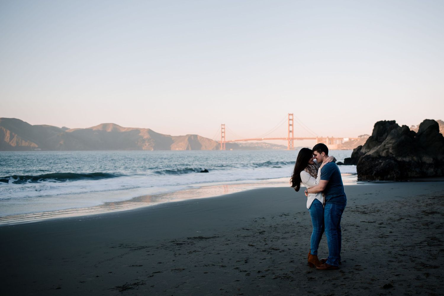 A couple embraces on a beach with the Golden Gate Bridge visible in the distance.