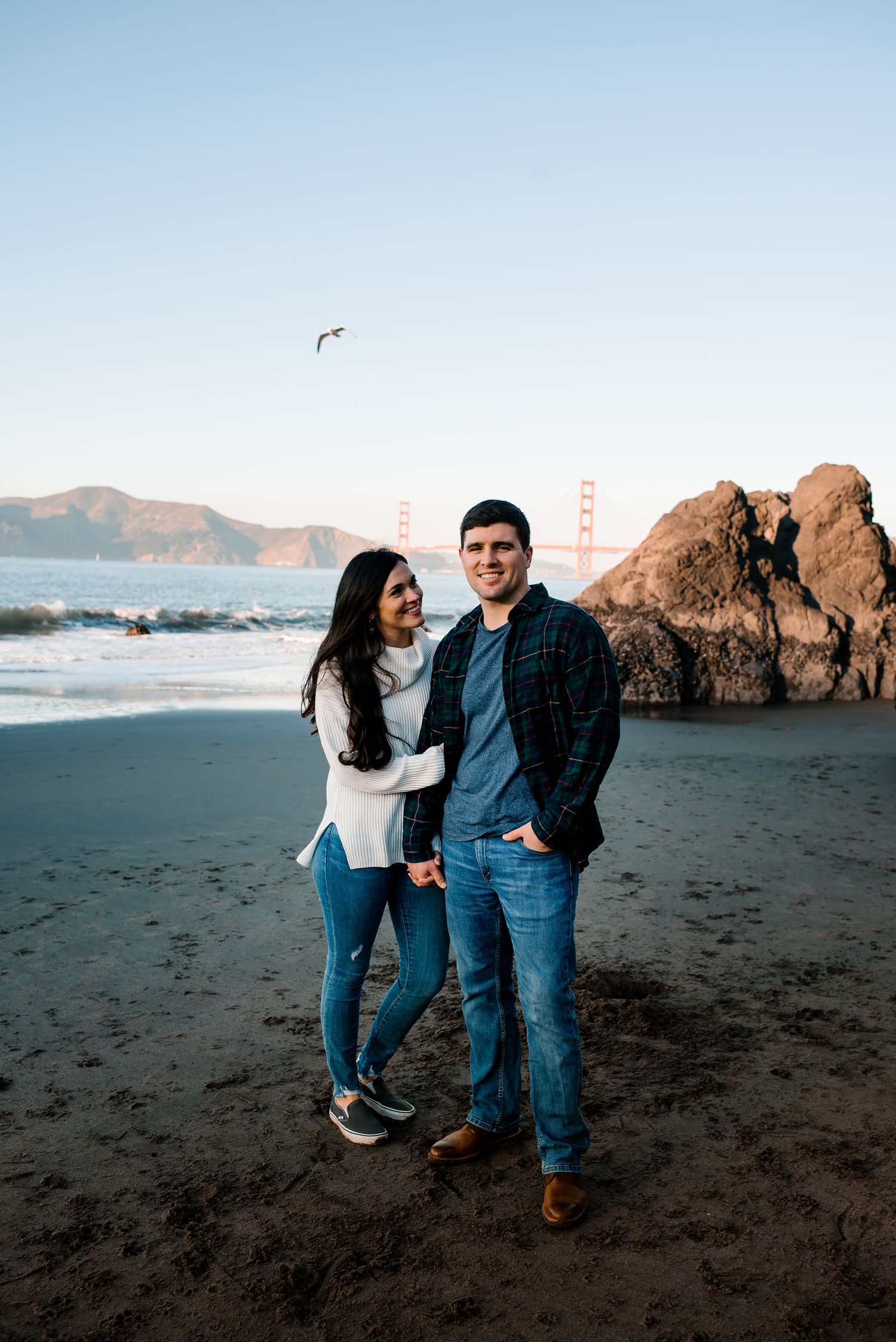 A couple walks along a rocky beach with the Golden Gate Bridge visible in the distance.