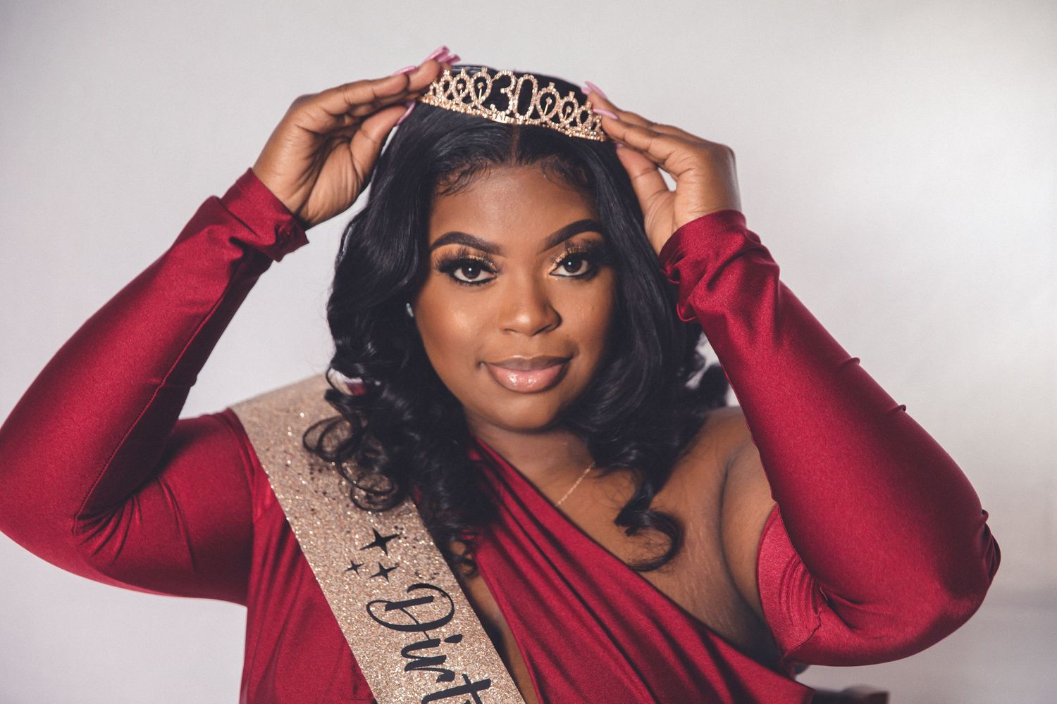 Young Black woman in red dress placing a crown on her head while wearing a birthday queen sash.