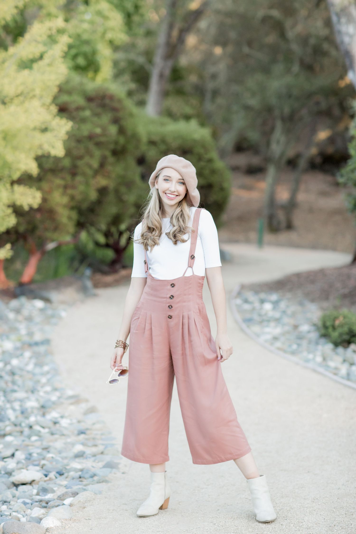 Young woman smiling in pink suspender culottes, white top, and blush beret on a garden path.