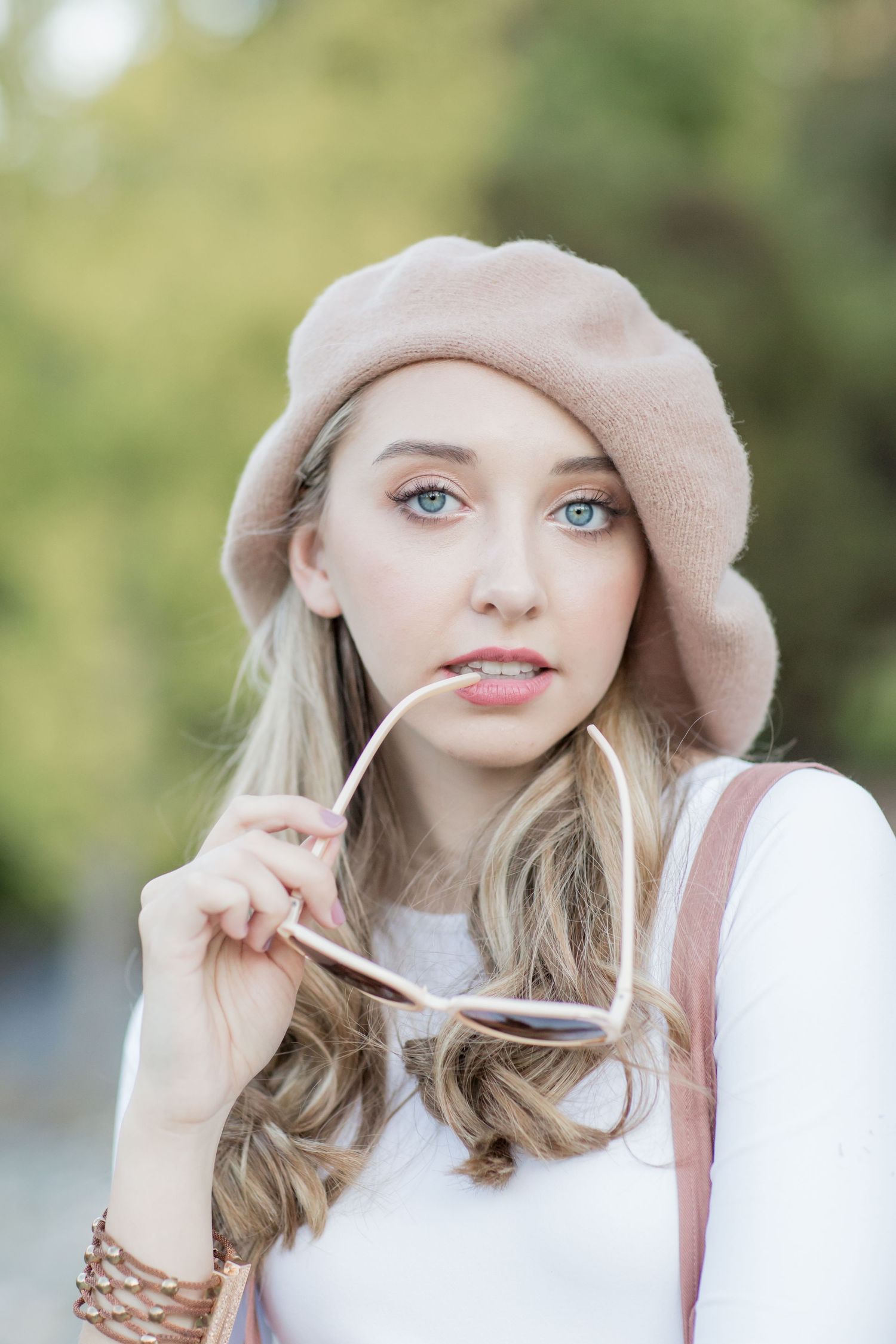 Close-up of blonde woman in blush beret holding sunglasses with blue eyes and soft makeup outdoors.