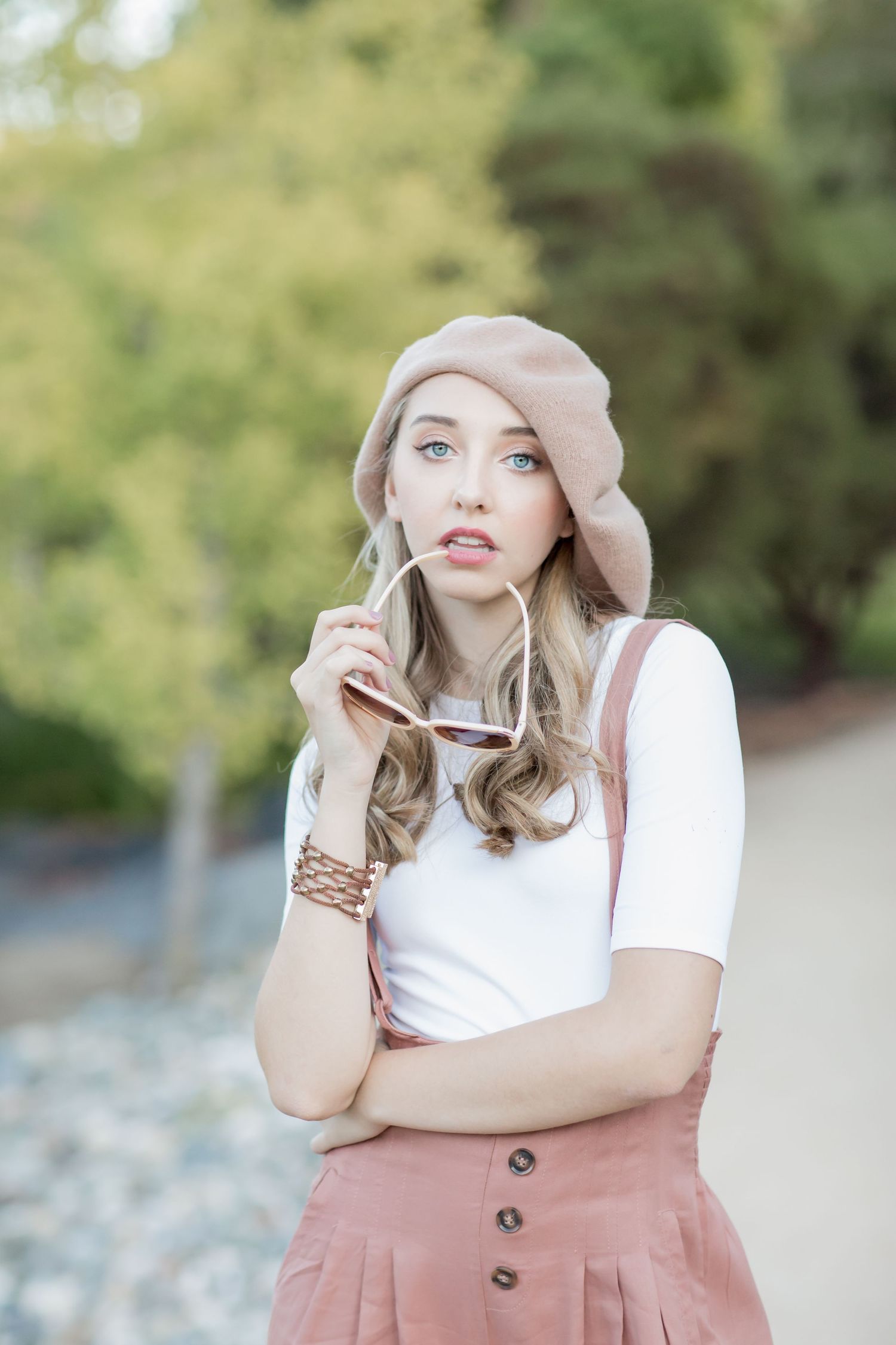 Blonde woman in pink beret holding sunglasses, wearing white top and pink suspender skirt outdoors.