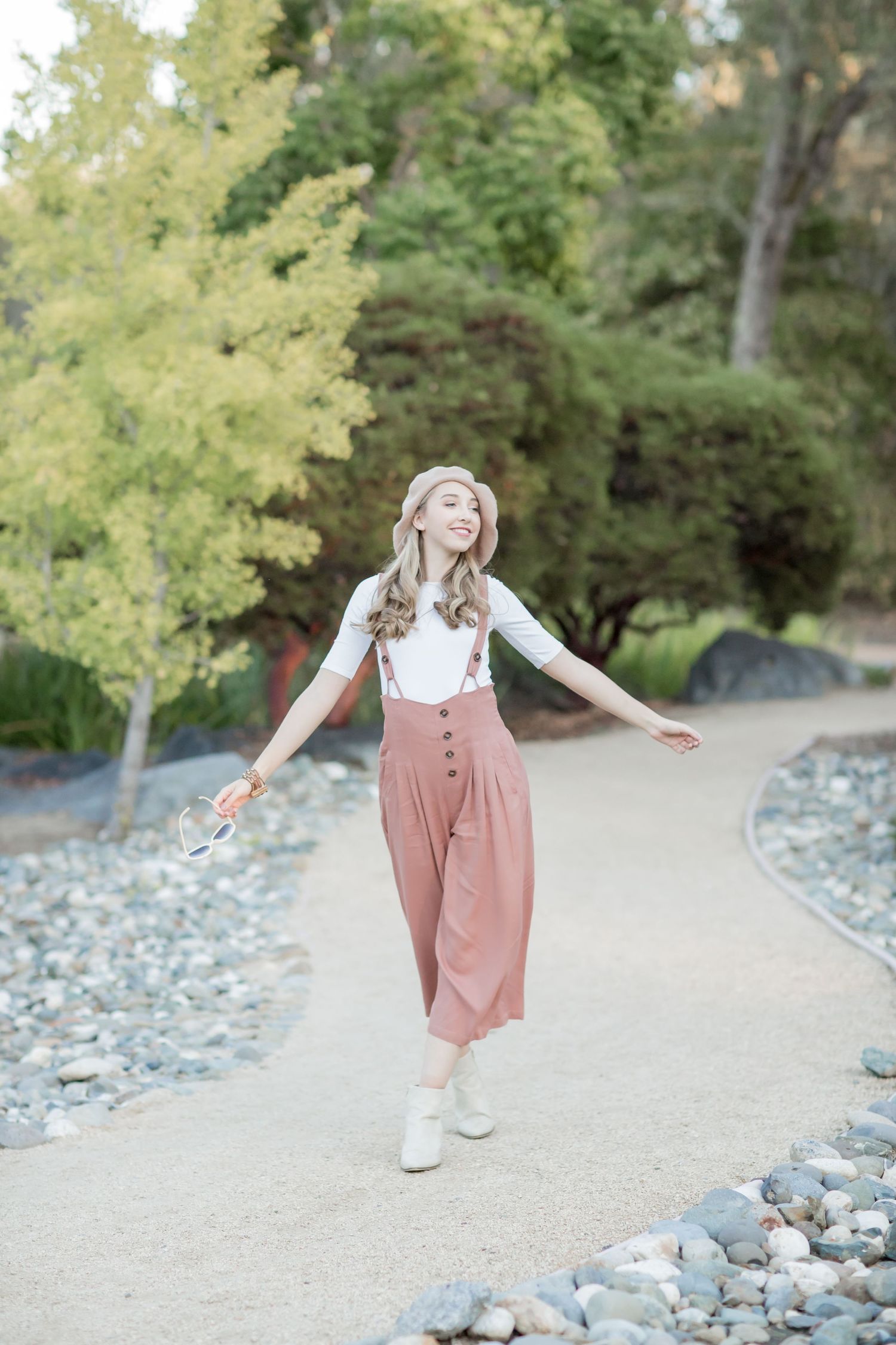 Joyful woman in pink beret and rust culottes twirling on a garden stone path outdoors.
