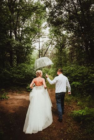 A couple walking along a wooded path while holding a clear umbrella.