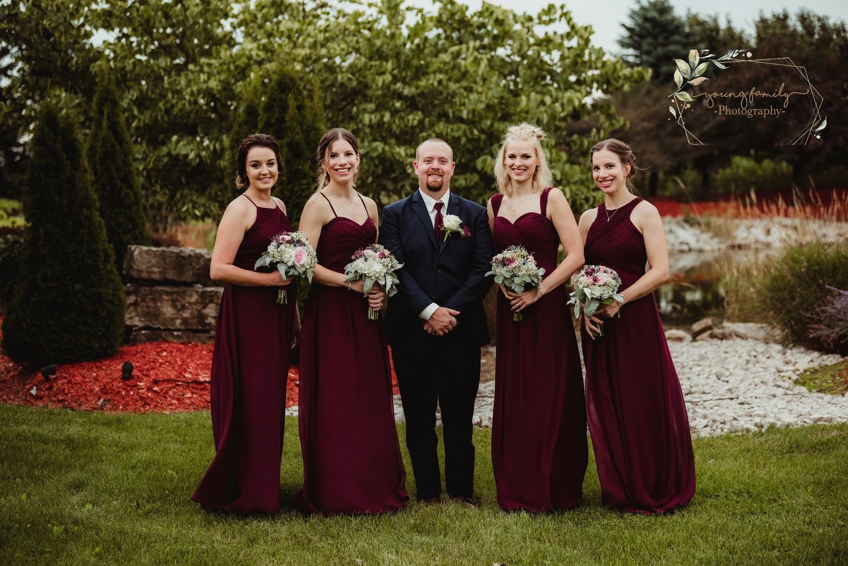 Wedding party in burgundy dresses poses outdoors holding white floral bouquets.