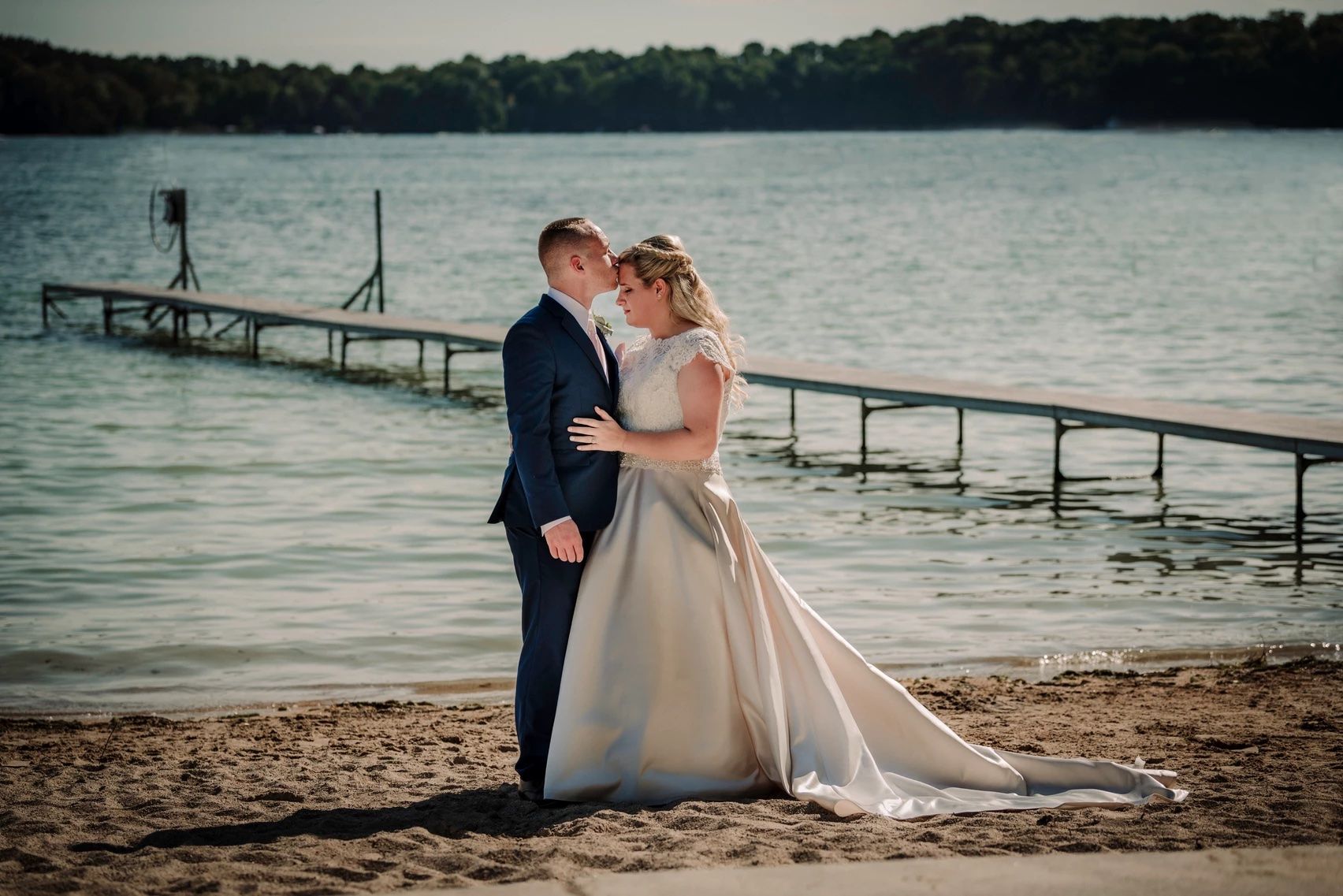 Newlyweds share romantic moments on a lakeside beach with wooden docks in the background.