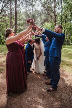 Wedding party members in burgundy and navy blue celebrate with raised arms in a forest setting.