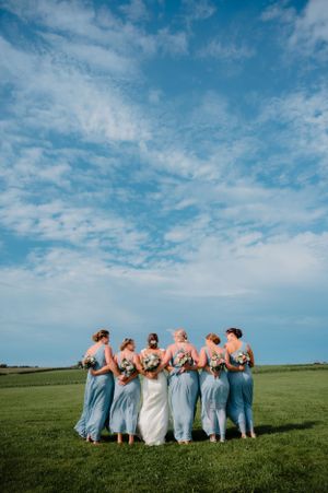 Wedding party in light blue attire posing together on a hilltop under bright blue sky.