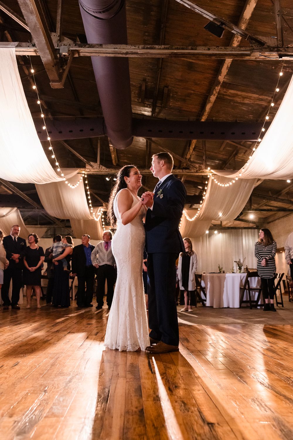 Couples dancing on a polished wooden floor in an elegant venue with white fabric draping and warm lighting.