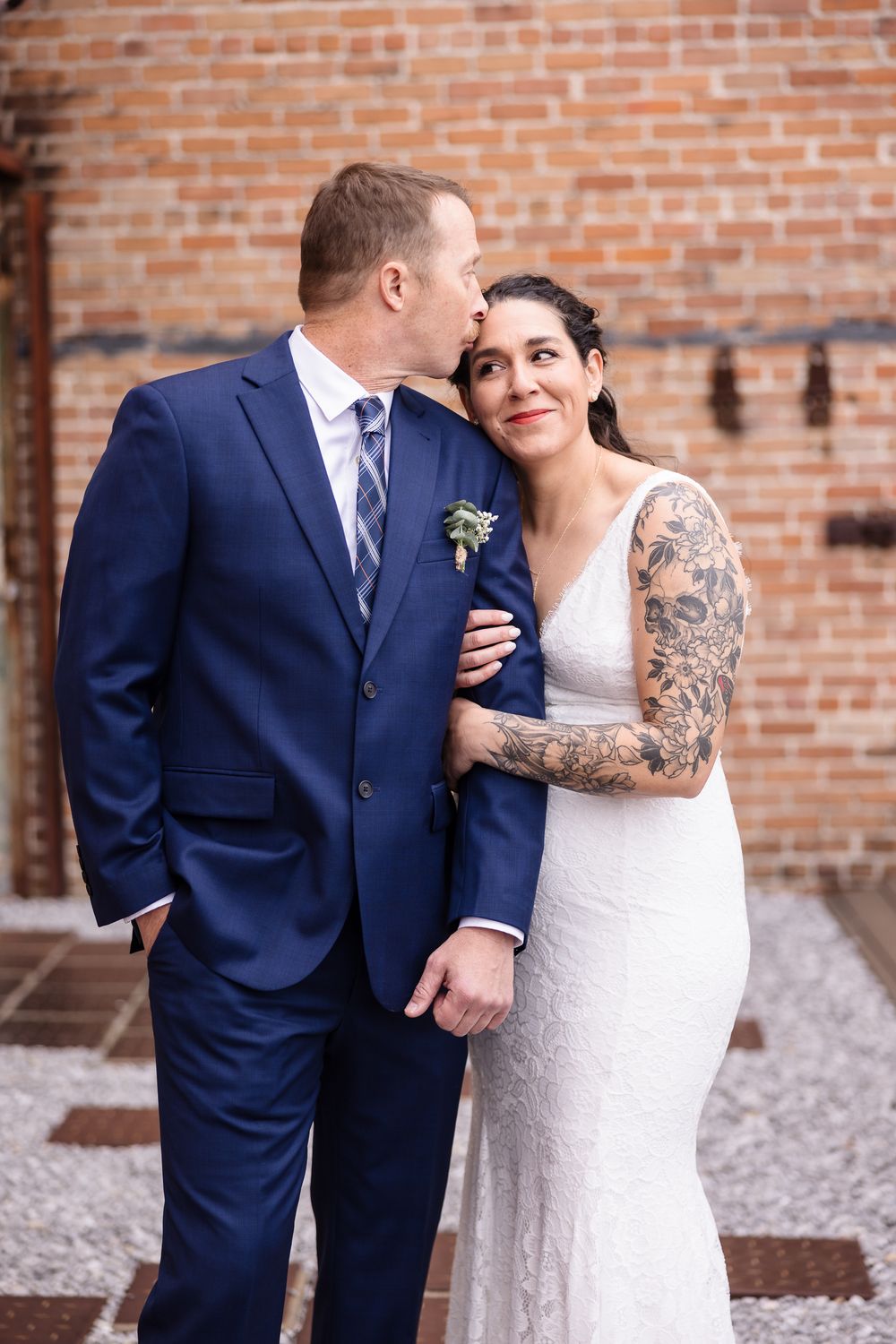 Wedding couple in navy suit and white dress pose against brick wall backdrop.