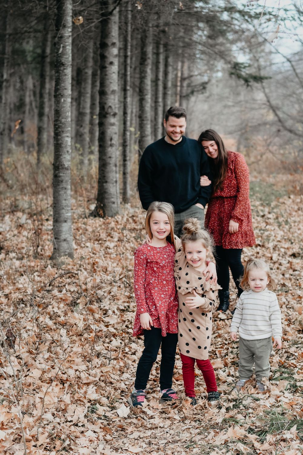 Family in coordinated red and neutral outfits poses among tall forest trees in autumn.