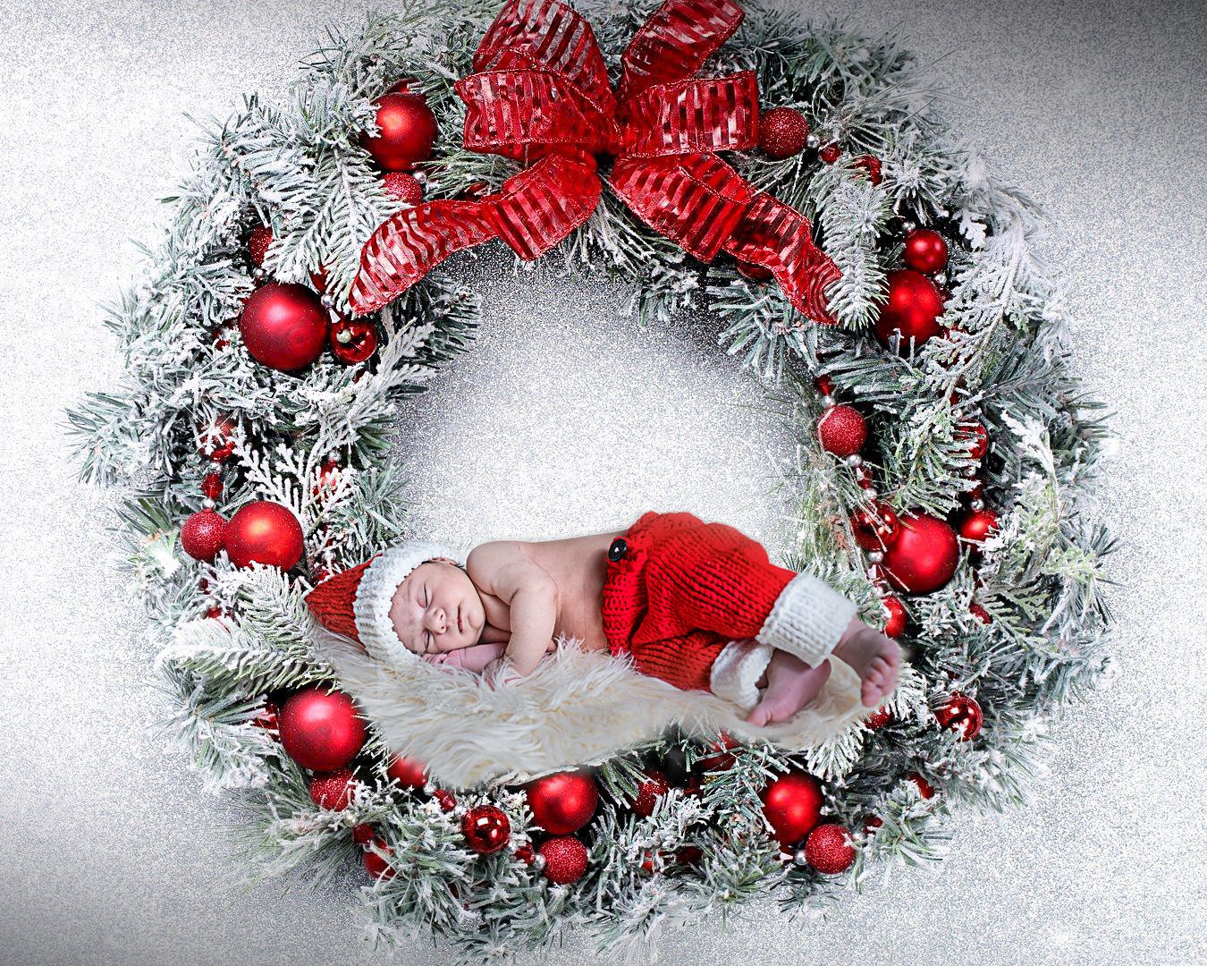 Baby dressed in Santa outfit sleeping inside festive Christmas wreath with red ornaments.