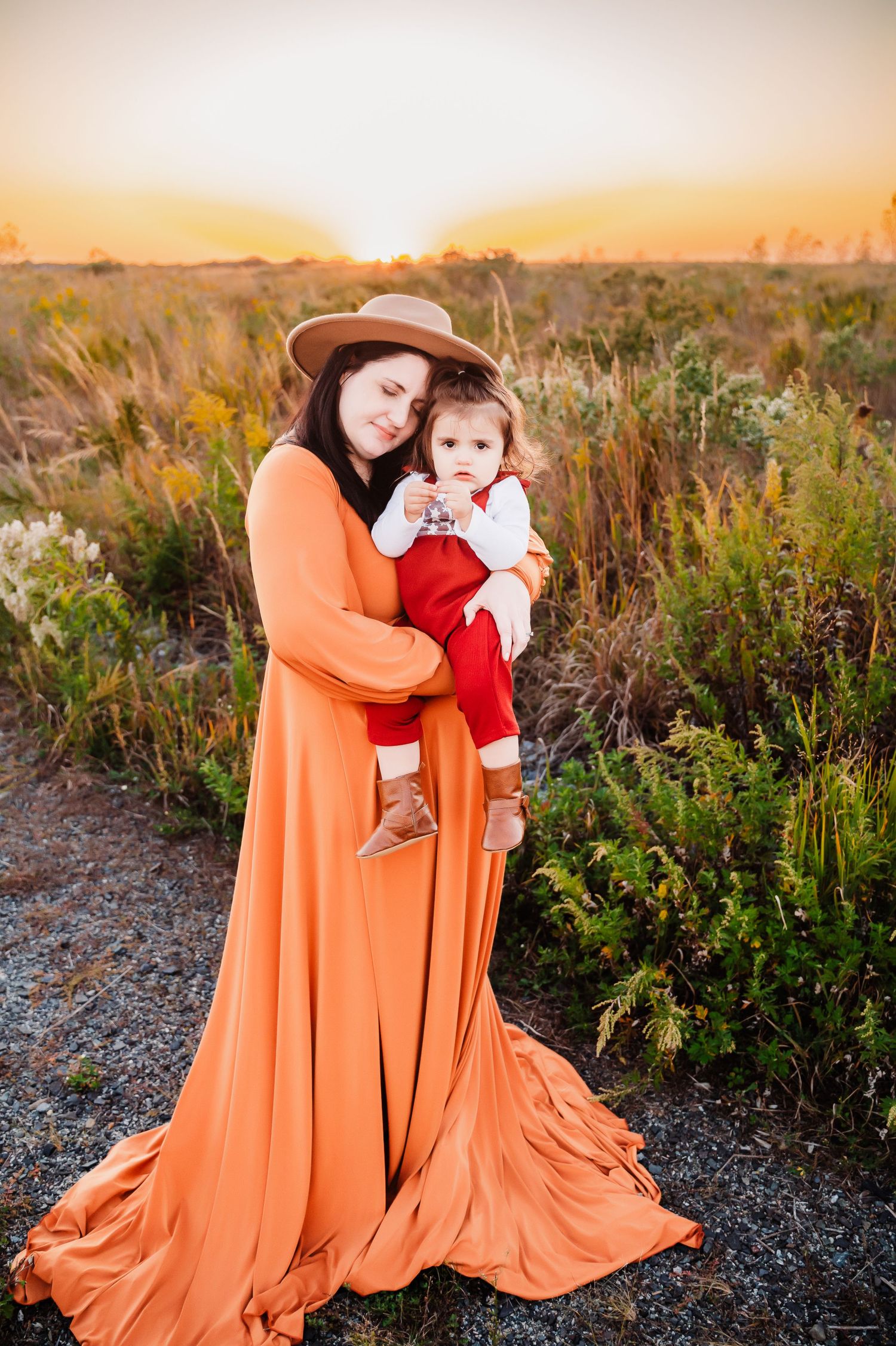 Mother wearing rust gown holding young daughter wearing red in a field in Staten Island, NY.