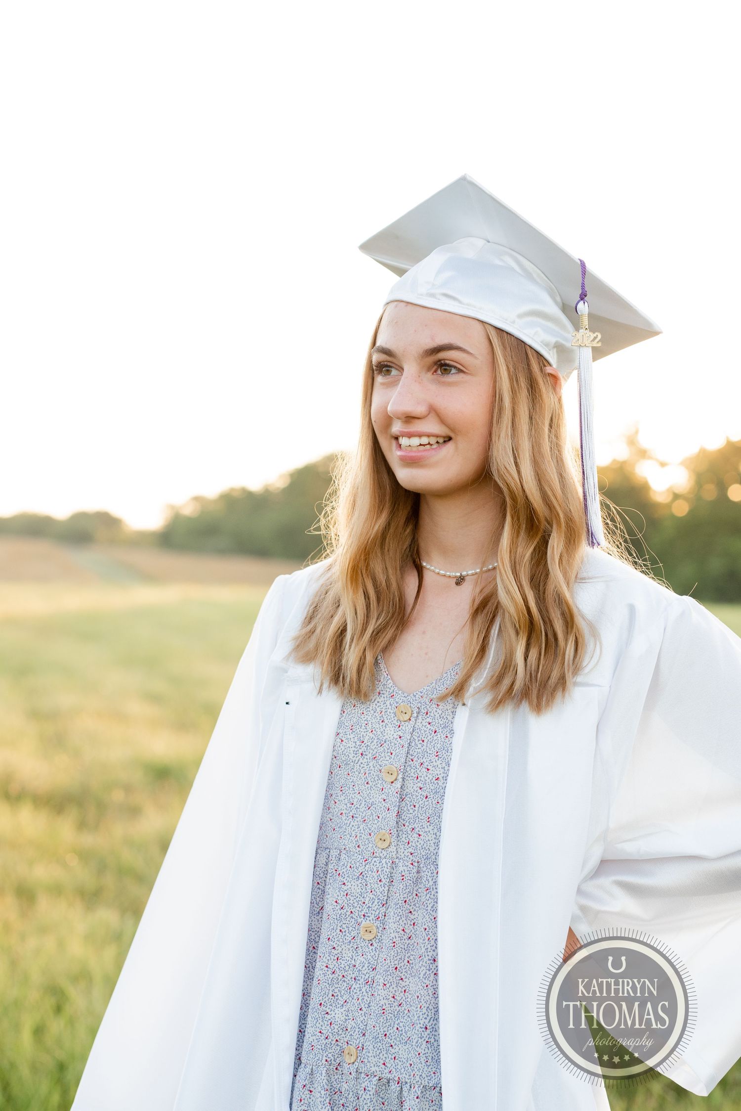 Graduate in white cap and gown smiles against golden sunset in field.