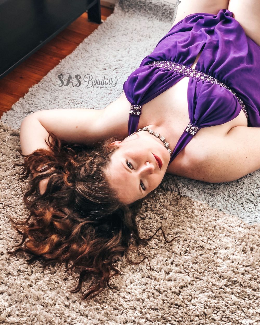 A person in a purple dress lies on a beige carpet with long curly brown hair spread out.