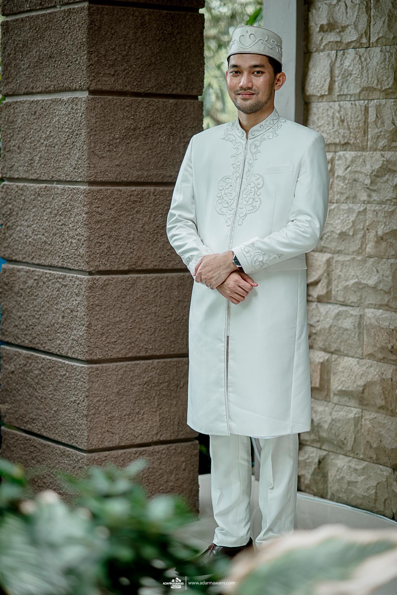 A groom in traditional white Islamic wedding attire poses against a stone wall.