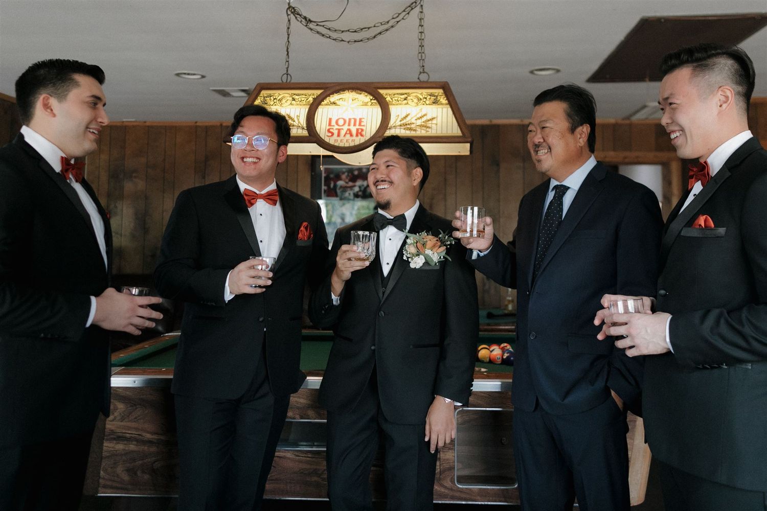 Five groomsmen in formal suits share drinks and conversation at a pre-wedding gathering.