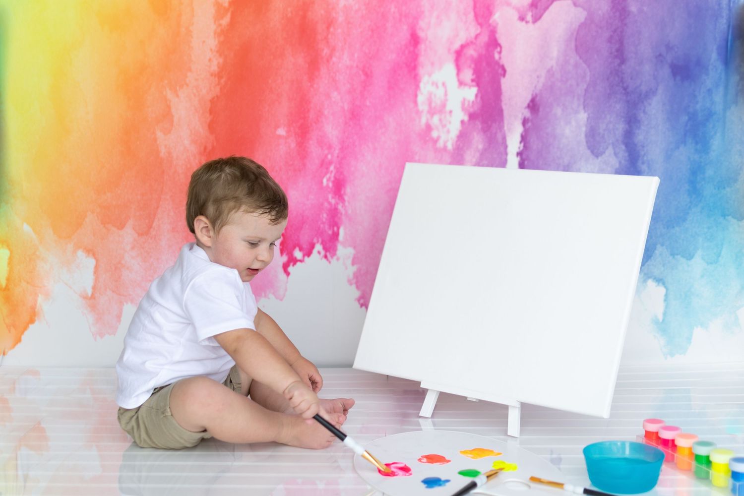 Young toddler painting on canvas easel against colorful rainbow watercolor backdrop.
