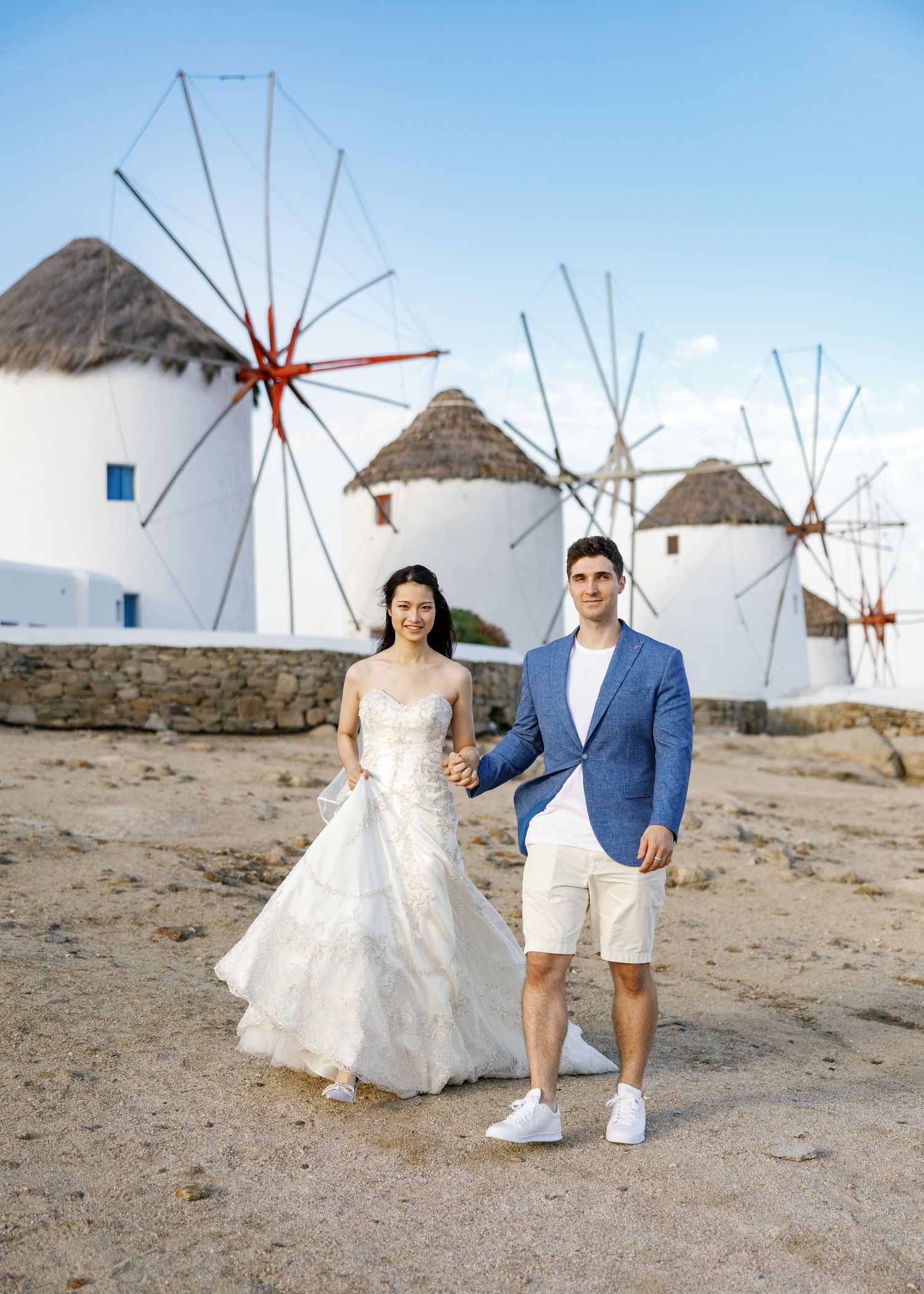 Couple in wedding attire poses in front of traditional white windmills in Mykonos, Greece.