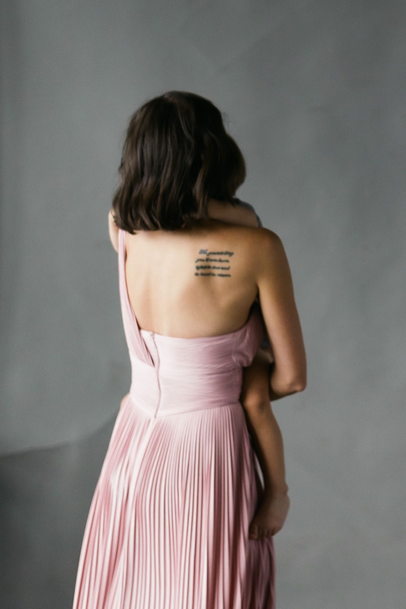 Back view of person wearing pink pleated dress with visible tattoo on upper back.