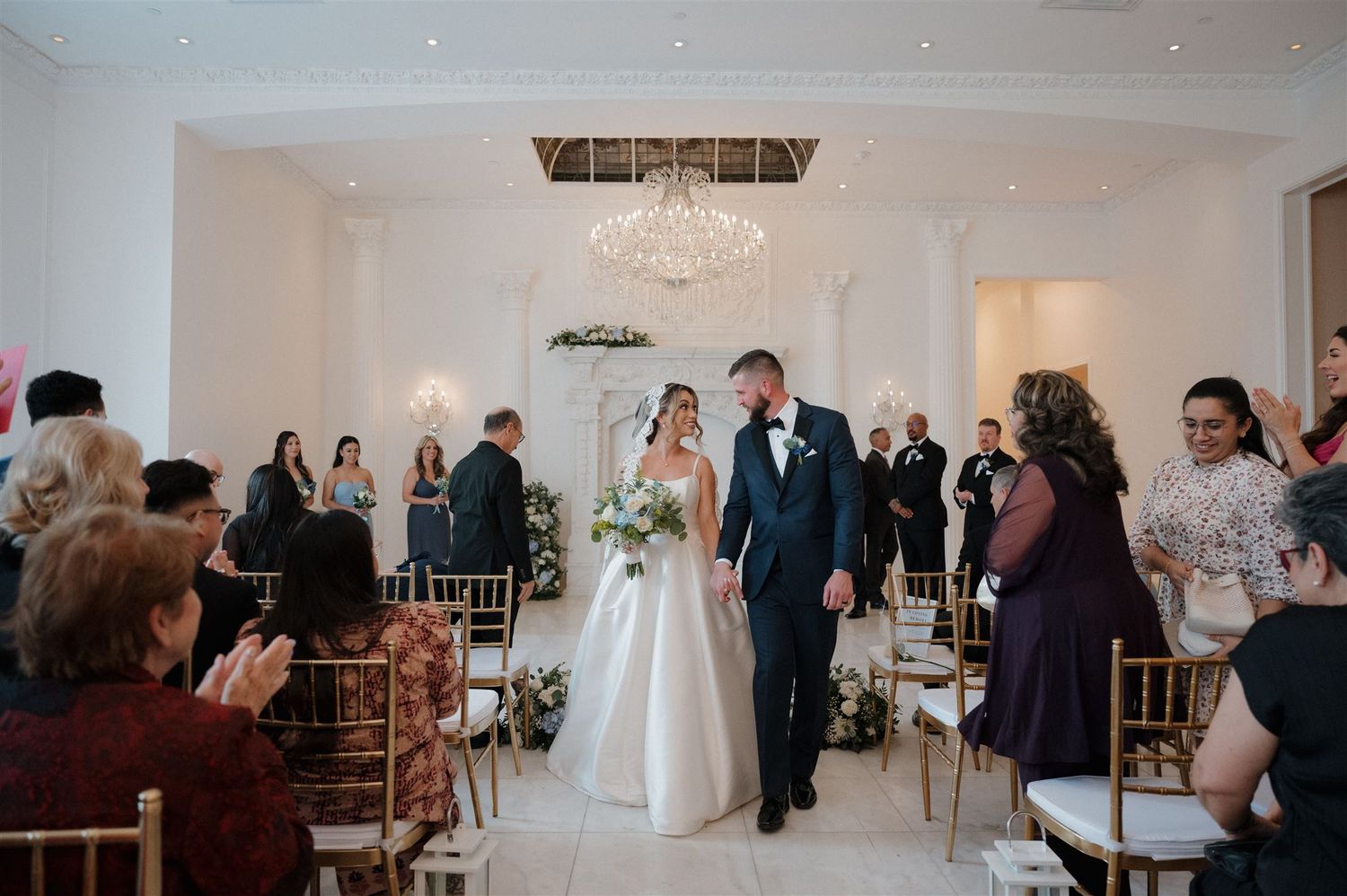A newly married couple walks down the aisle while wedding guests stand and applaud in an elegant white reception venue.