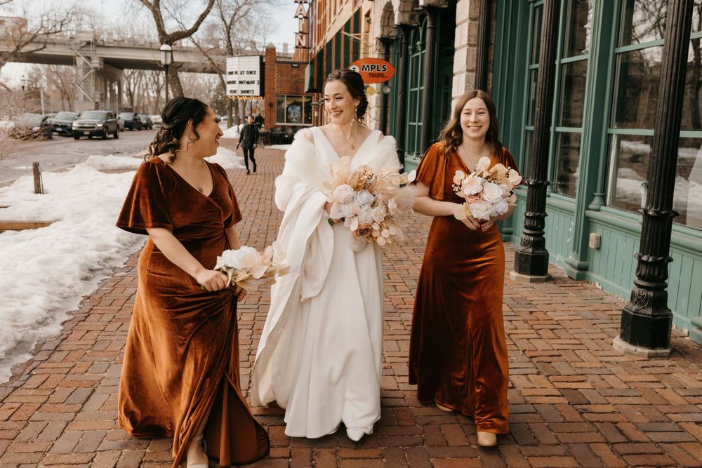 Bride and bridesmaids in rust velvet dresses walking on brick sidewalk in winter holding bouquets.