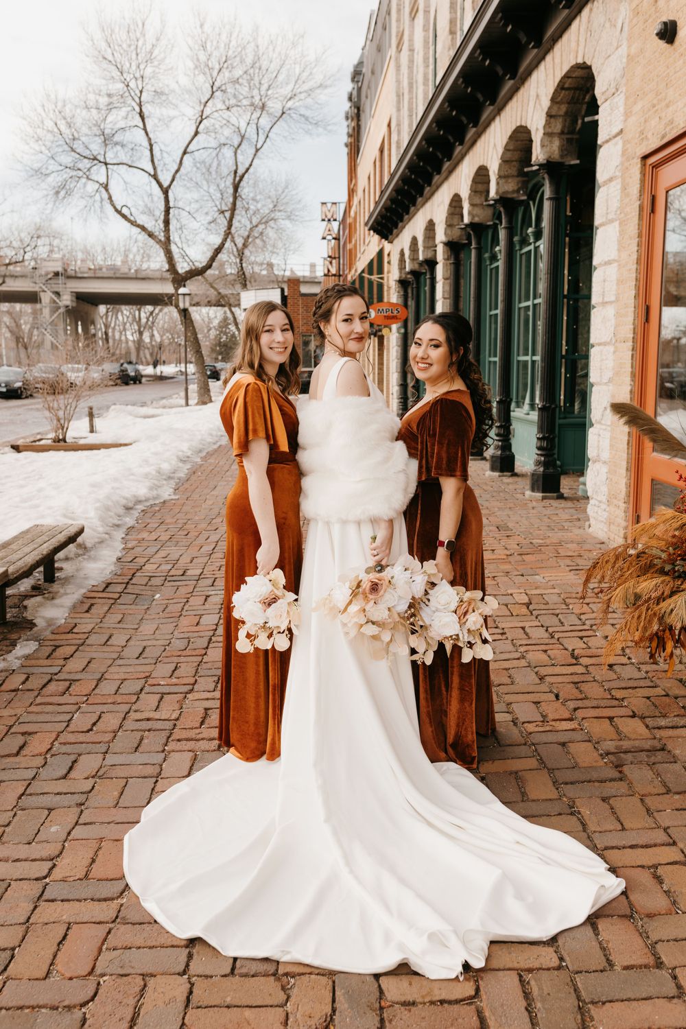 Bride with fur stole and two bridesmaids in rust velvet gowns posing on brick path in winter.