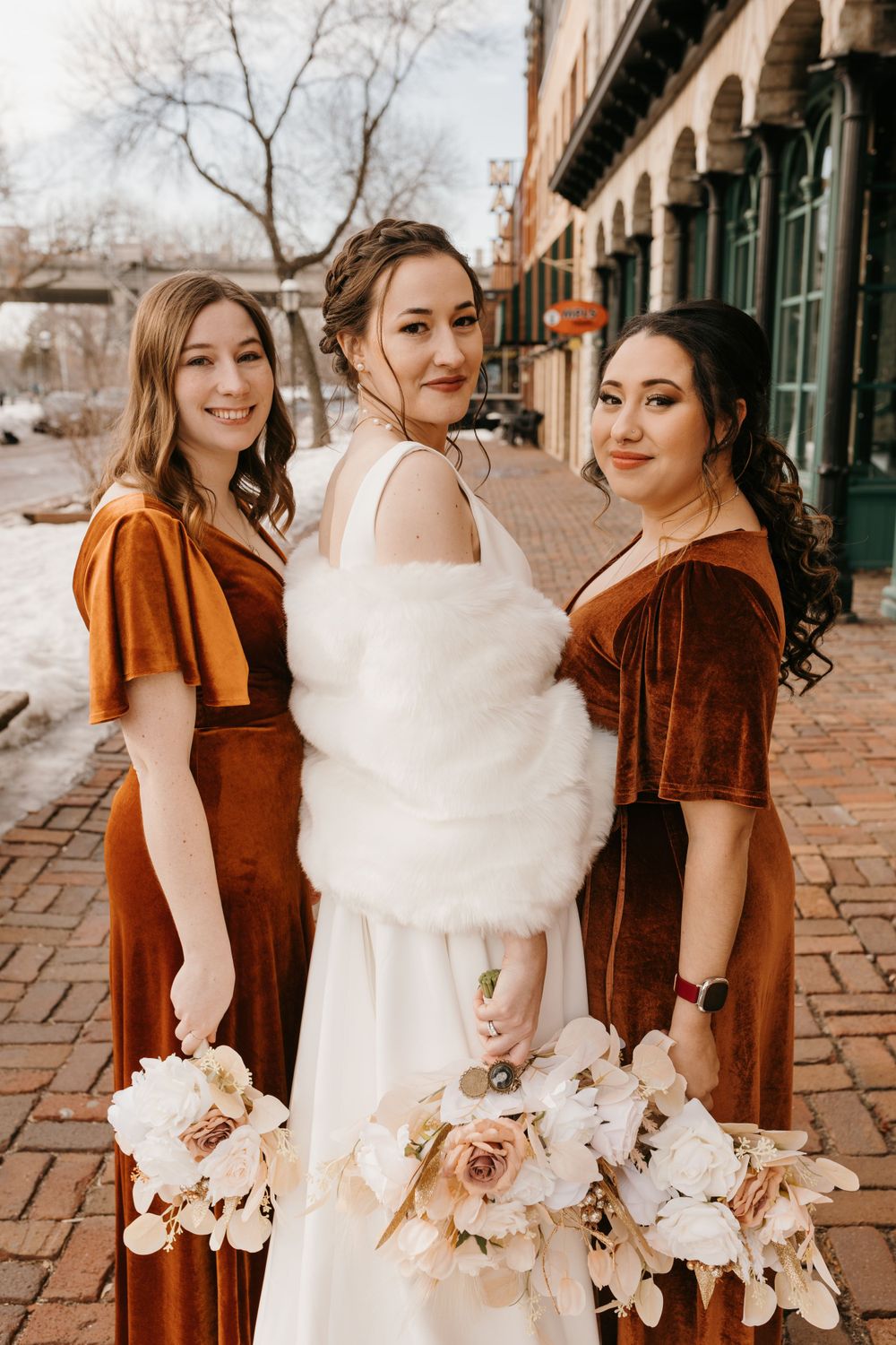 Bride wearing white fur wrap poses with two bridesmaids in copper velvet dresses holding bouquets.