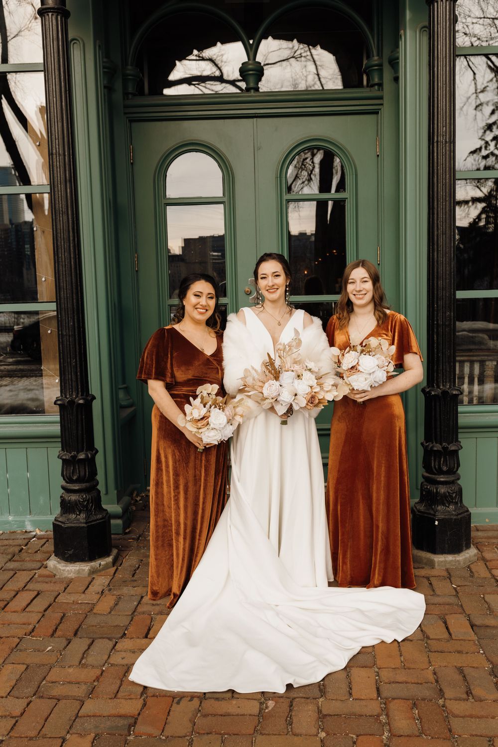 Bride in white gown with two bridesmaids in rust velvet dresses holding bouquets before green door.