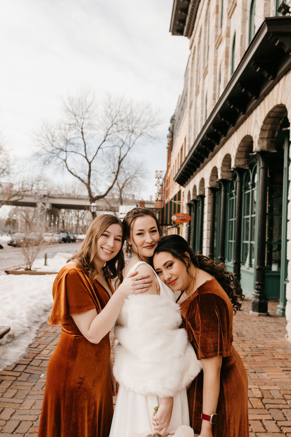 Two bridesmaids in rust velvet dresses hugging smiling bride in white fur wrap outdoors in winter.