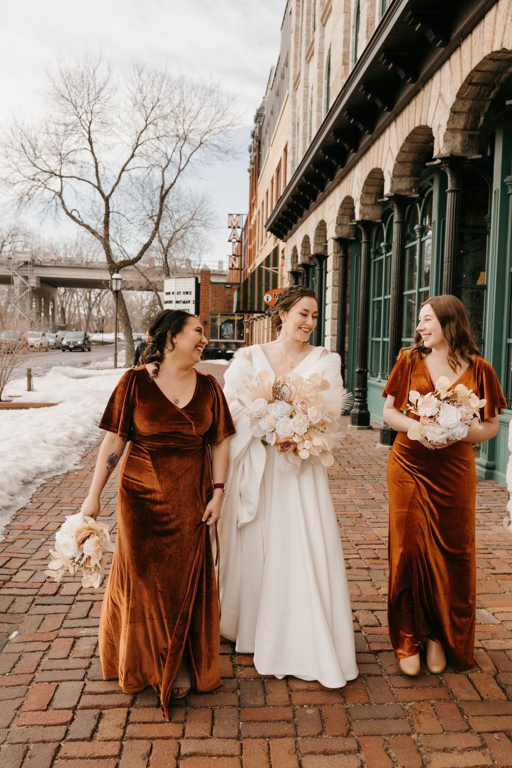 Bride in white gown with fur wrap walks with bridesmaids in burnt orange velvet dresses outdoors.
