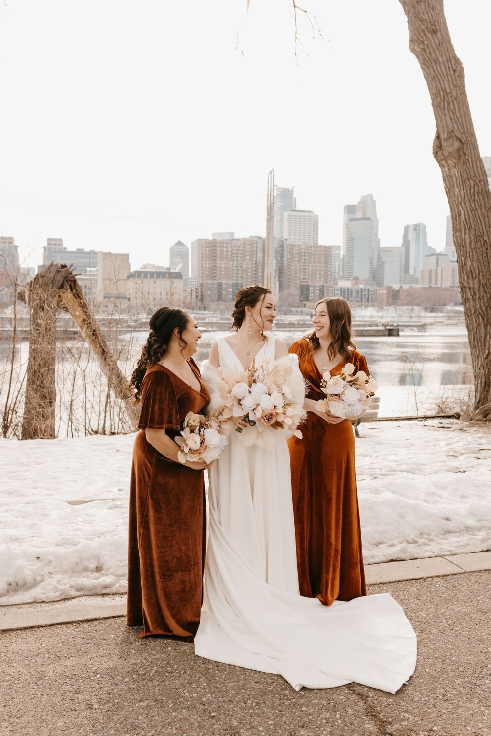 Bride and two bridesmaids in burnt orange velvet gowns holding bouquets with city skyline backdrop.