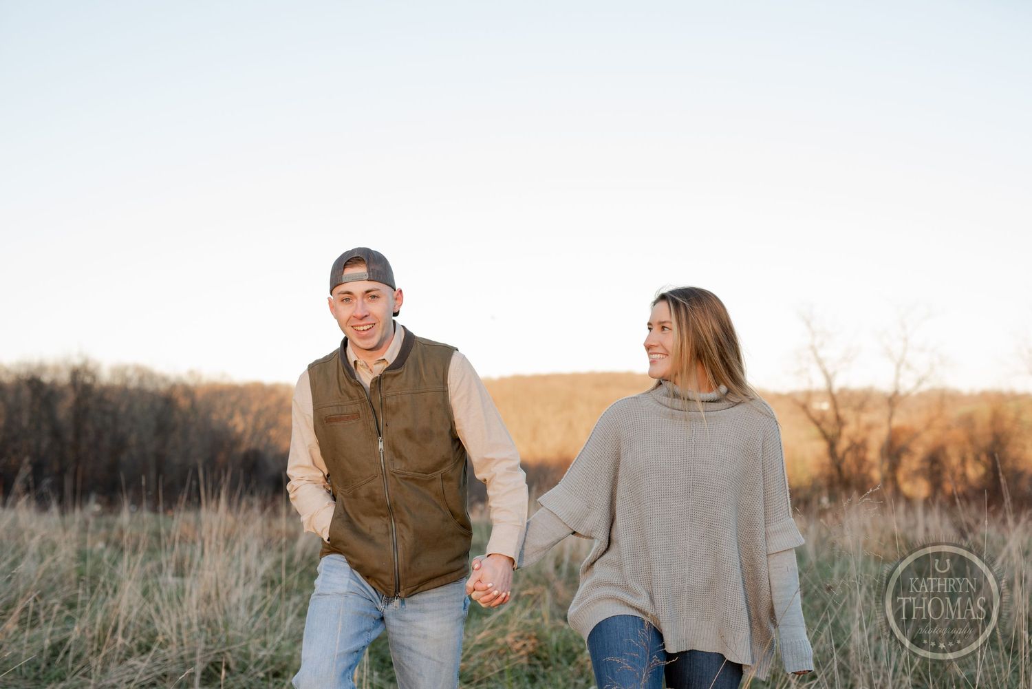 A couple walks hand in hand through a rustic field during a golden sunset photoshoot.