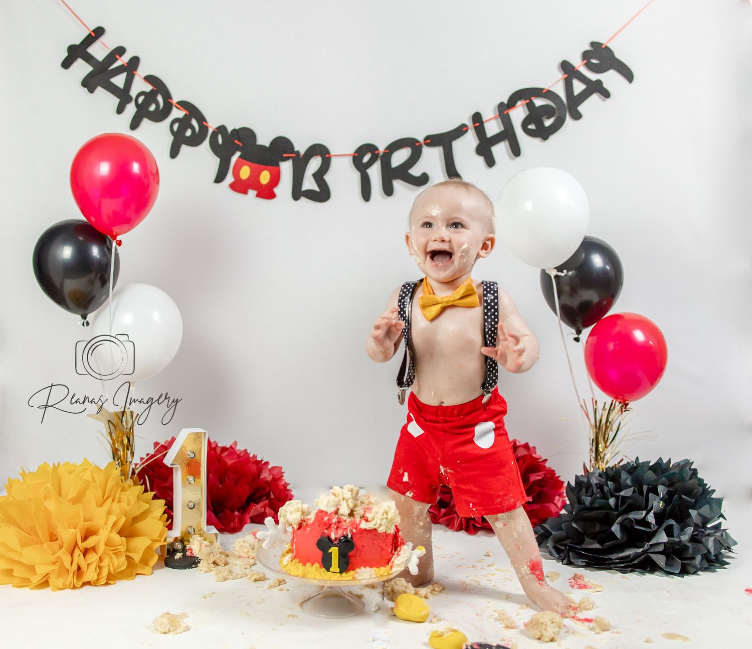 Excited baby boy in Mickey Mouse themed outfit celebrating first birthday smash cake party.