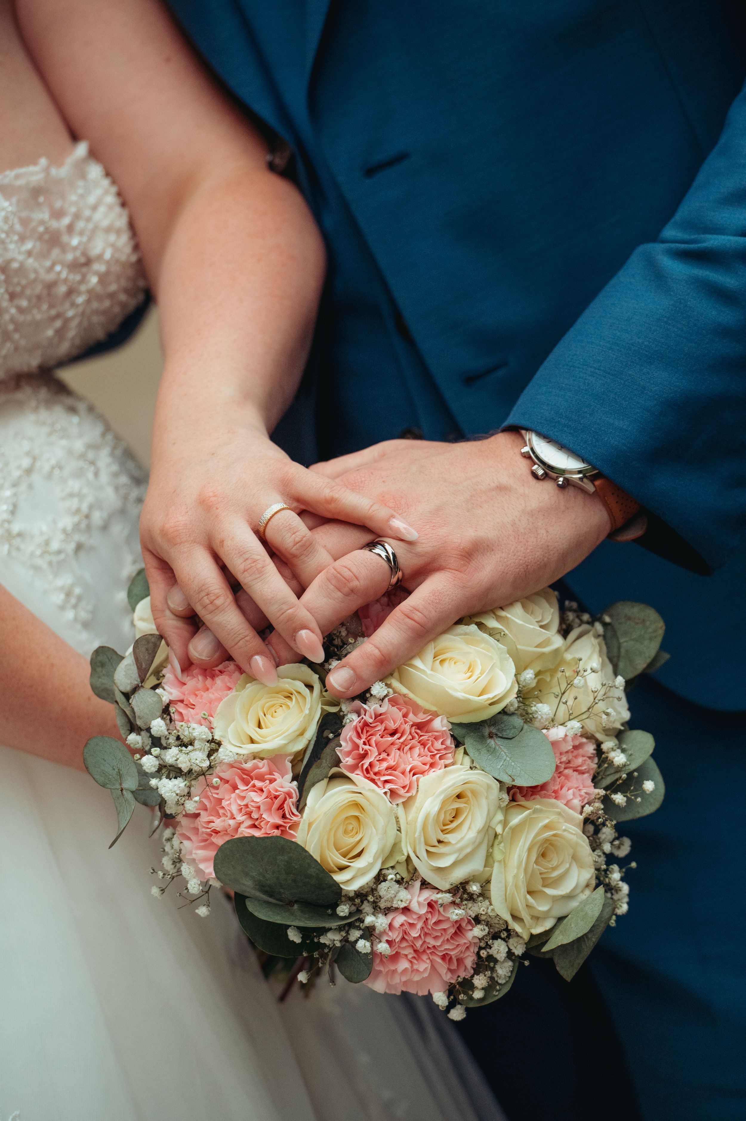Newlyweds display wedding rings on hands resting over a bridal bouquet of pink carnations and white roses.