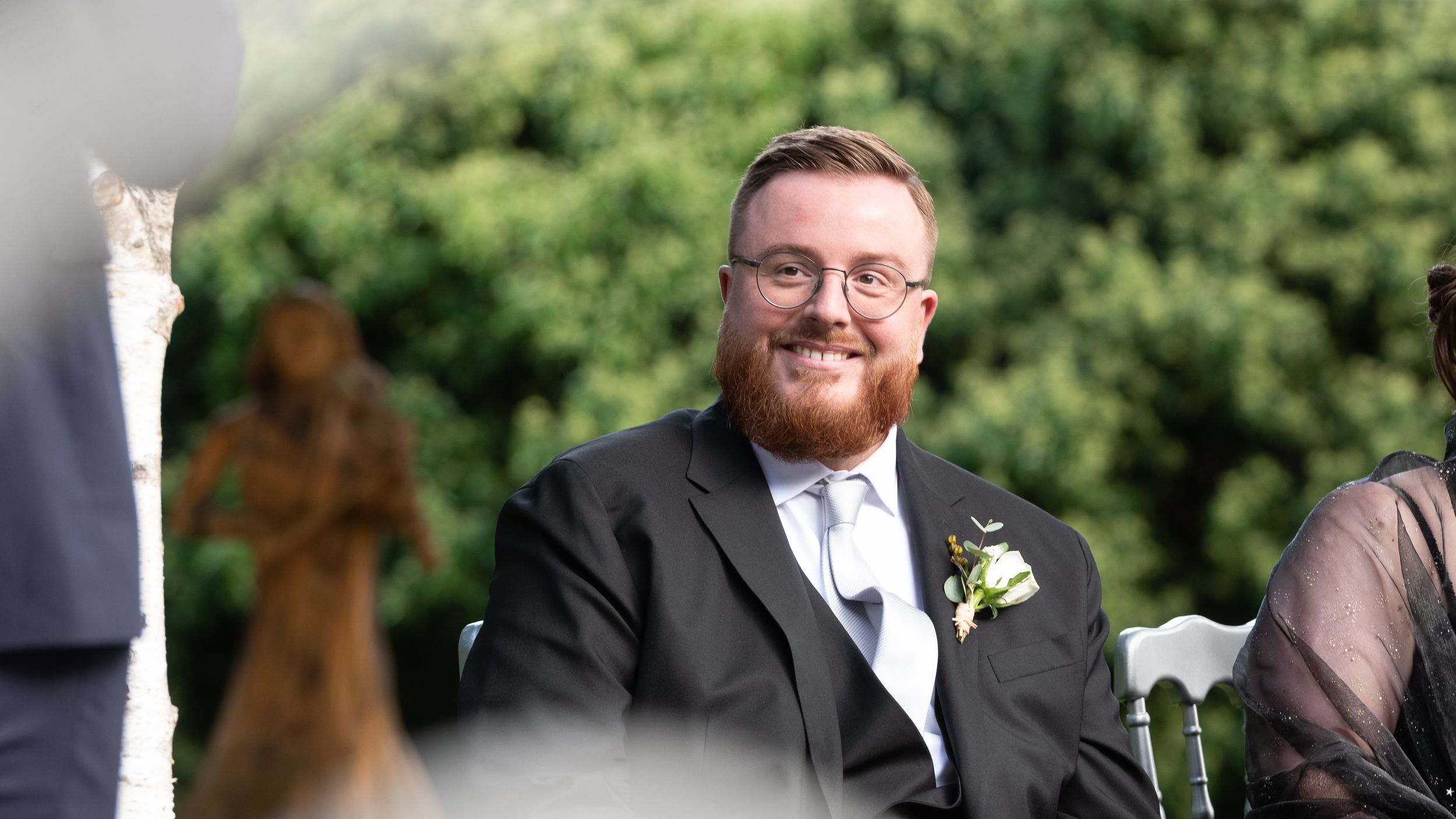 A smiling bearded man in a black suit with boutonniere enjoys an outdoor wedding ceremony.
