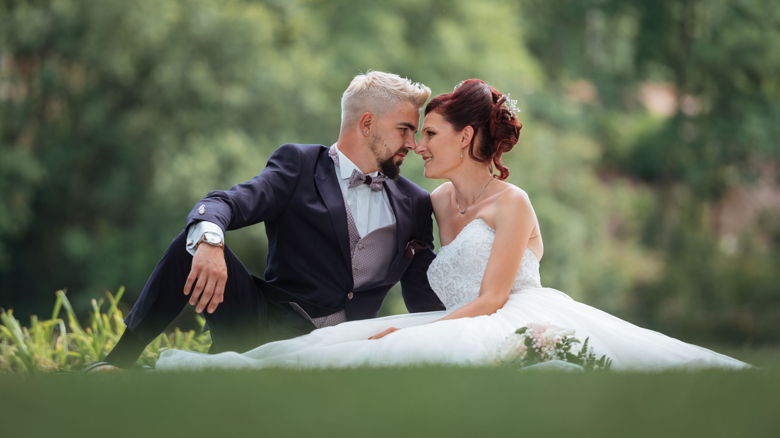 A bride and groom sit intimately together on green grass in a lush outdoor wedding setting.