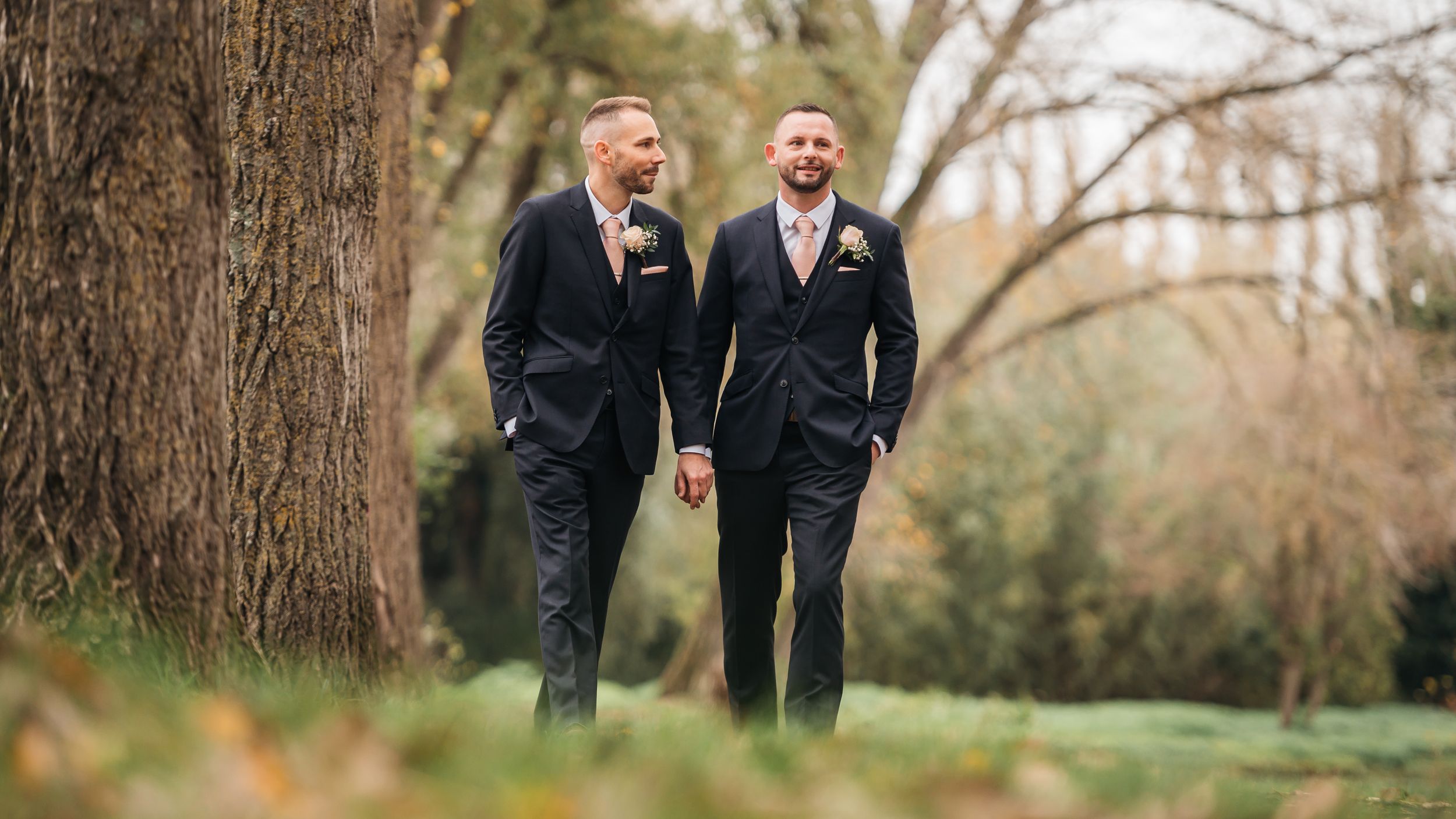 Two grooms in navy suits walk hand in hand through a tree-lined park on their wedding day.