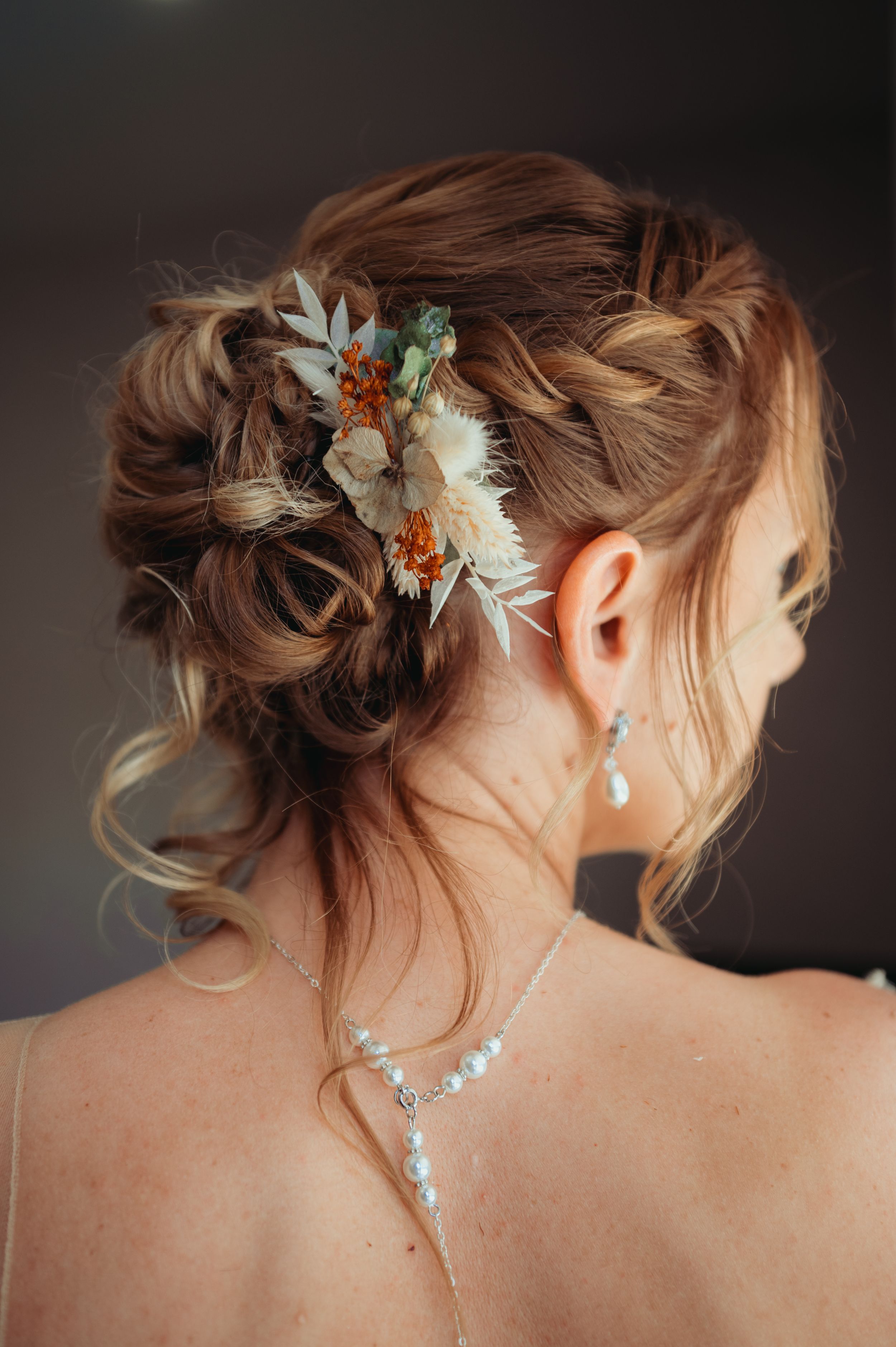 Elegant bridal updo with braids adorned with dried flowers and pearl drop earrings viewed from behind.