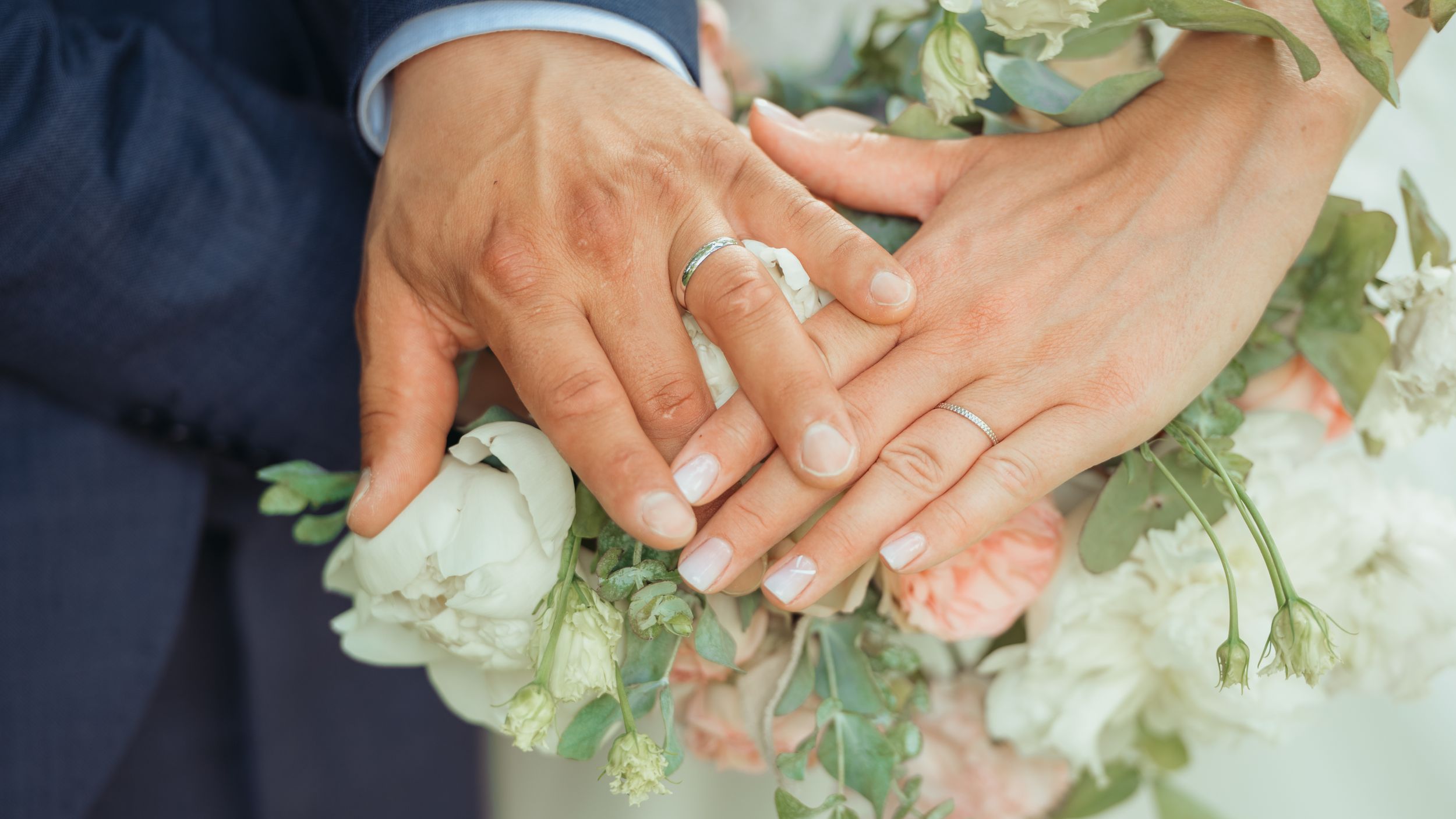 Bride and groom display wedding rings while hands rest over a bouquet of white and blush flowers.