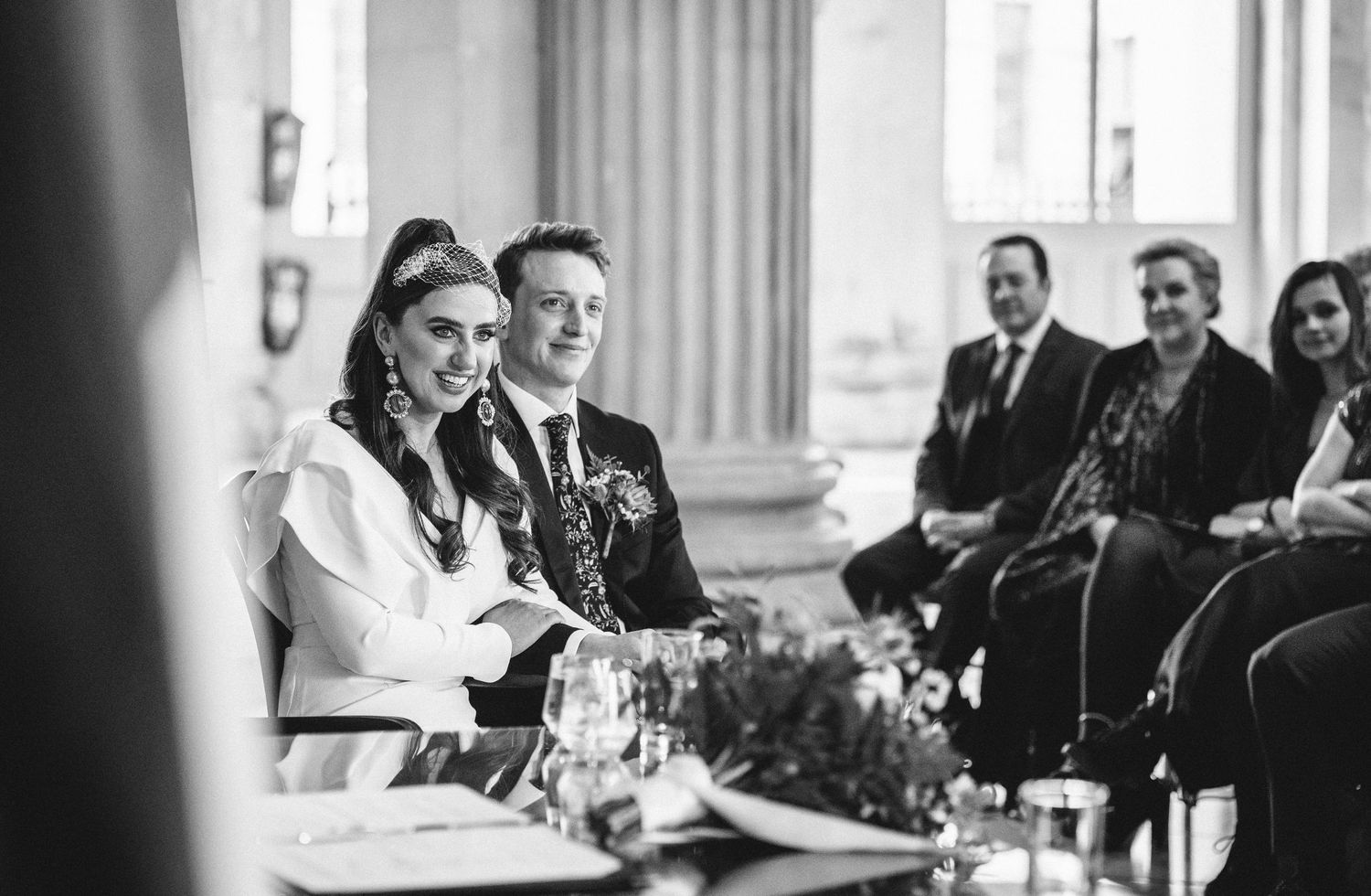 Candid black and white wedding reception photo showing newlyweds at a head table with guests seated in background.